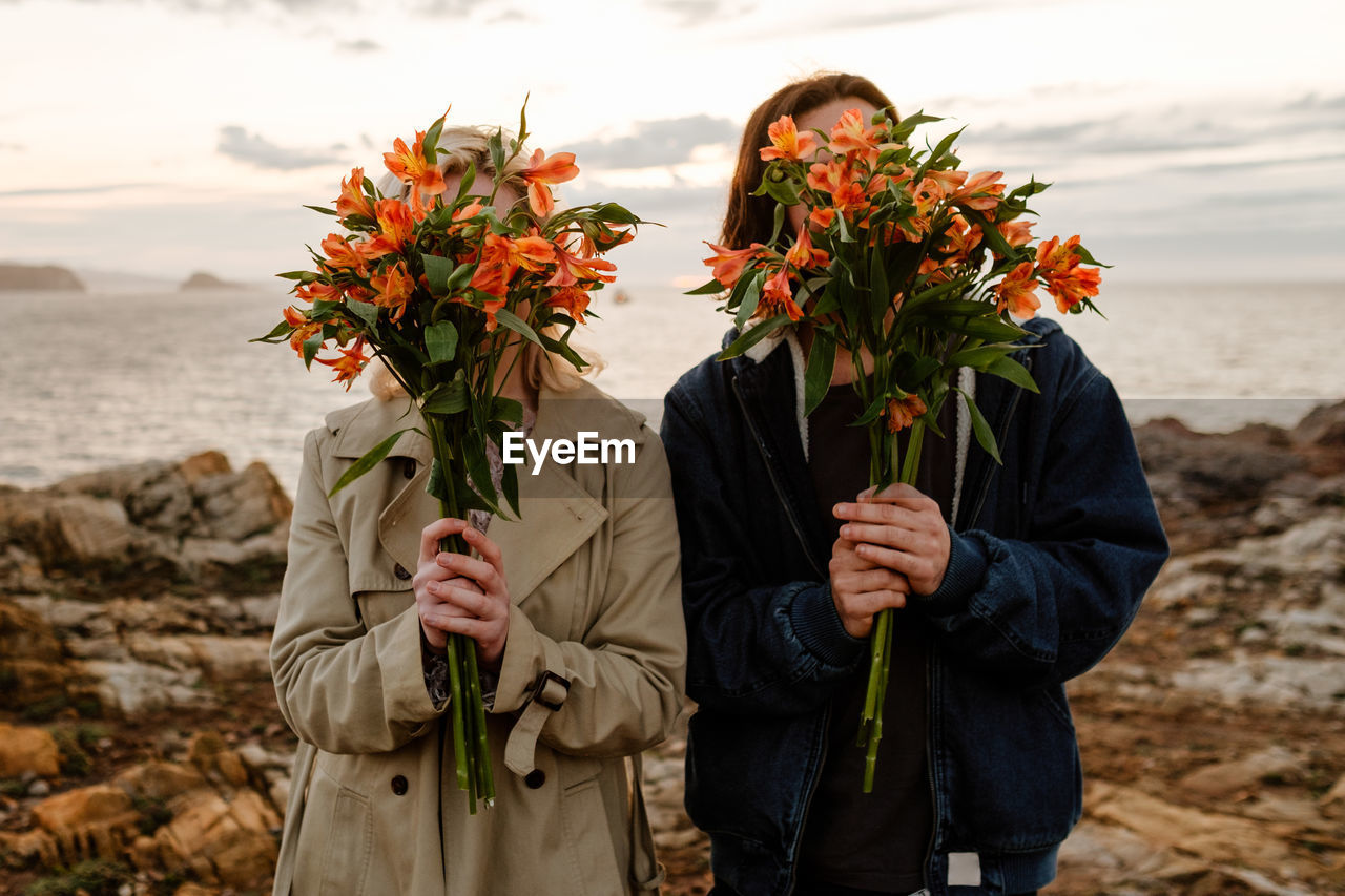 Anonymous boyfriend and girlfriend in outerwear covering faces with fresh bouquets while standing on seashore at sunset in aviles, spain