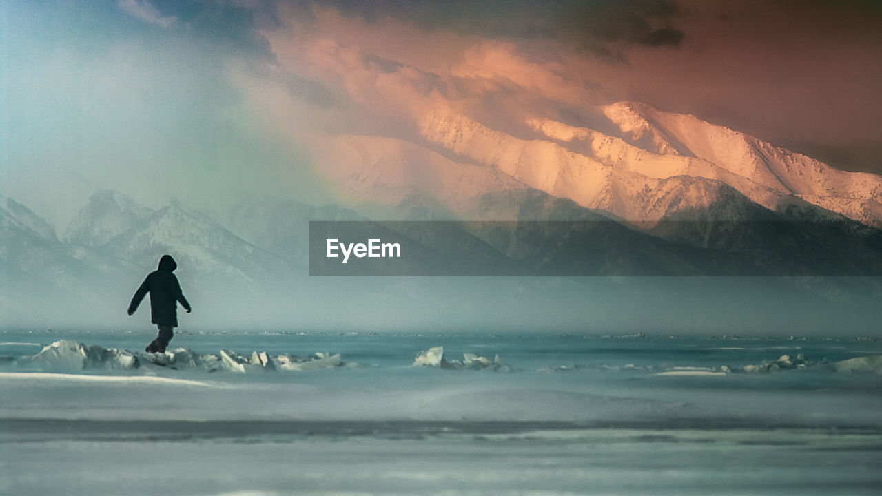 One person in windy cold weather at sunset on frozen lake with mountains at the background