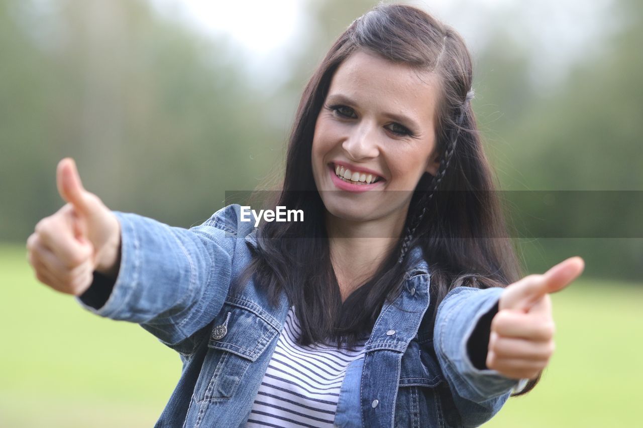 Portrait of smiling young woman gesturing thumbs up against trees