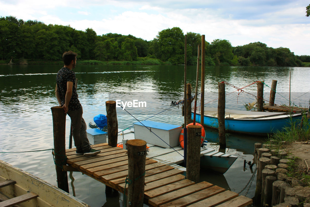 BOY STANDING ON PIER OVER LAKE AGAINST SKY