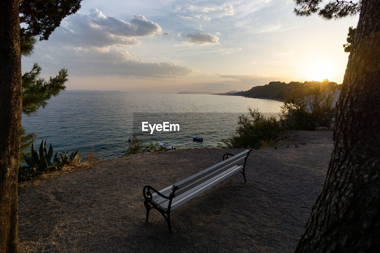 Empty bench by sea against sky during sunset