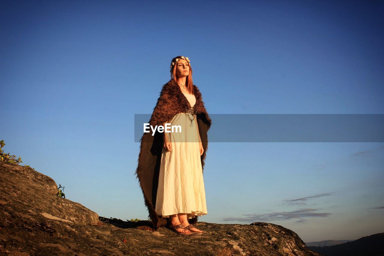 Low angle view of young woman standing on cliff against blue sky