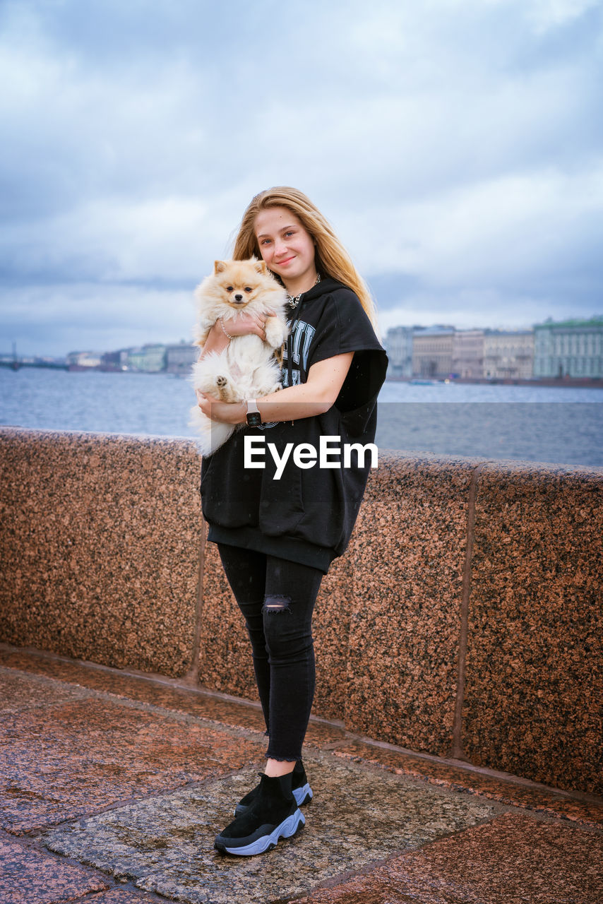 Cheerful and happy girl teenager holds and hugs pomeranian spitz on embankment