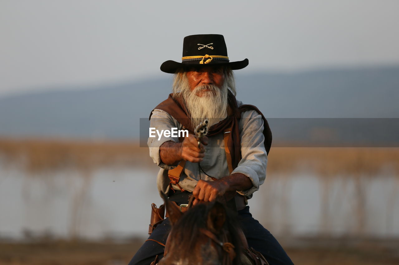 Portrait of man riding horse while holding handgun against sky
