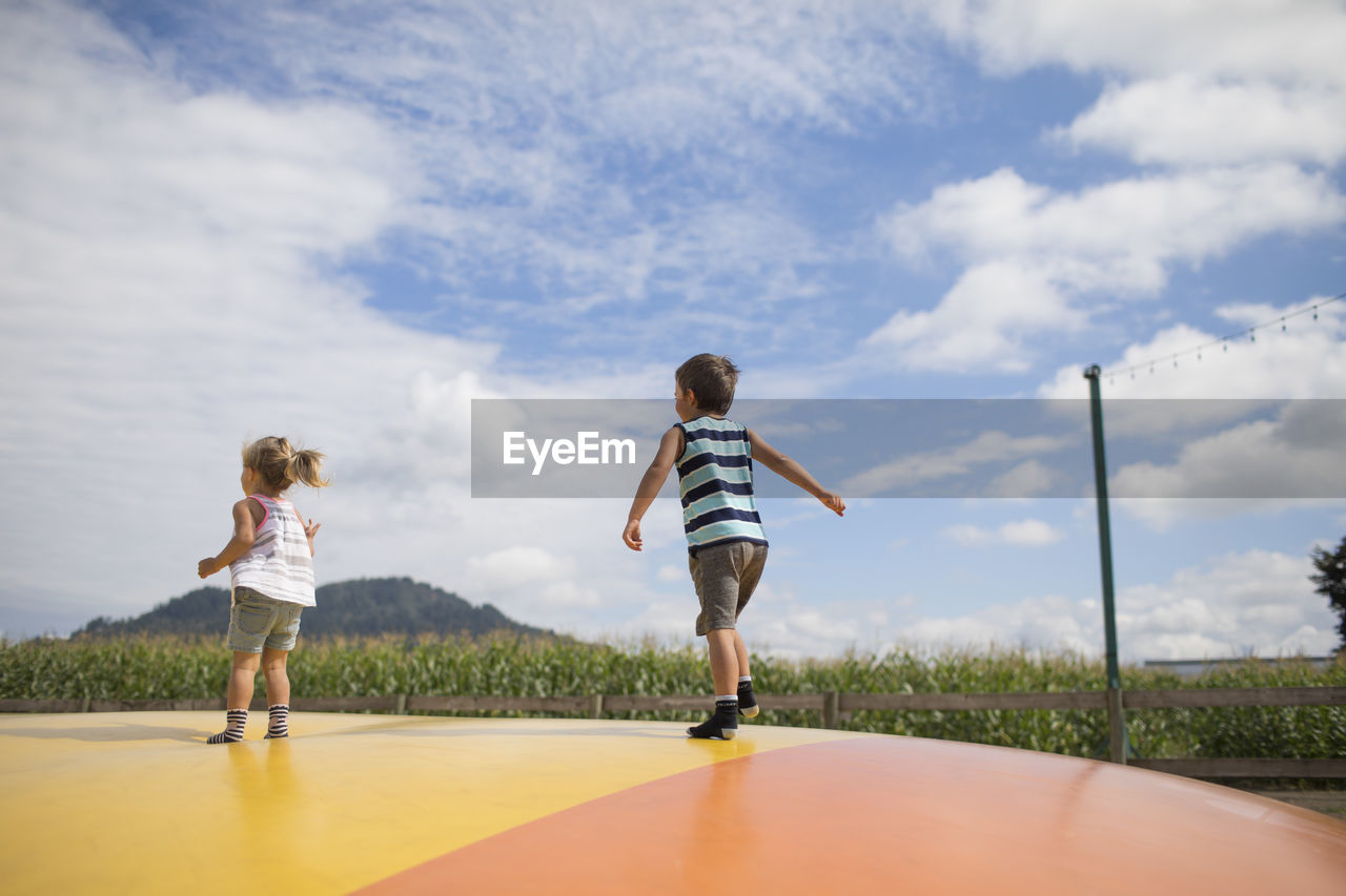 Rear view of boy and girl jumping on large trampoline outdoors.