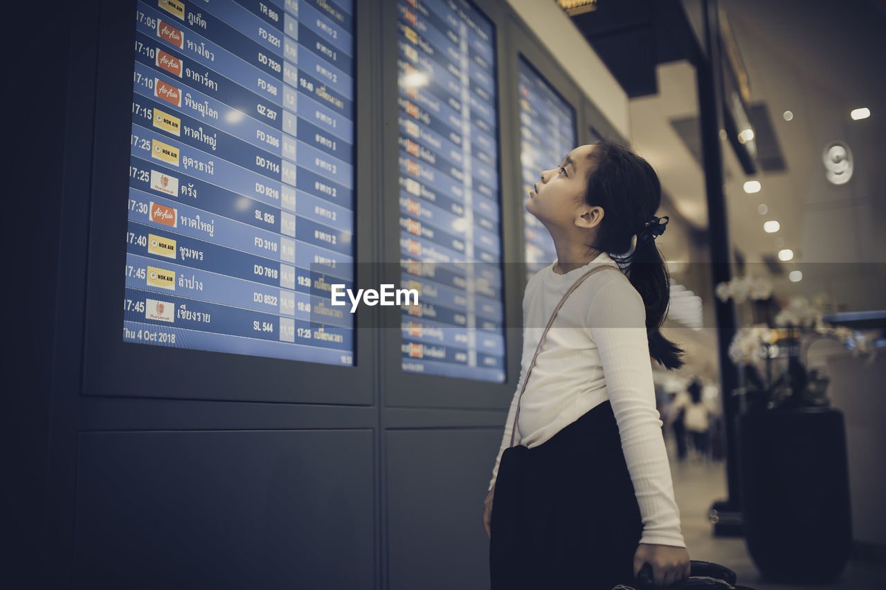 Cute girl looking arrival departure board while standing in airport