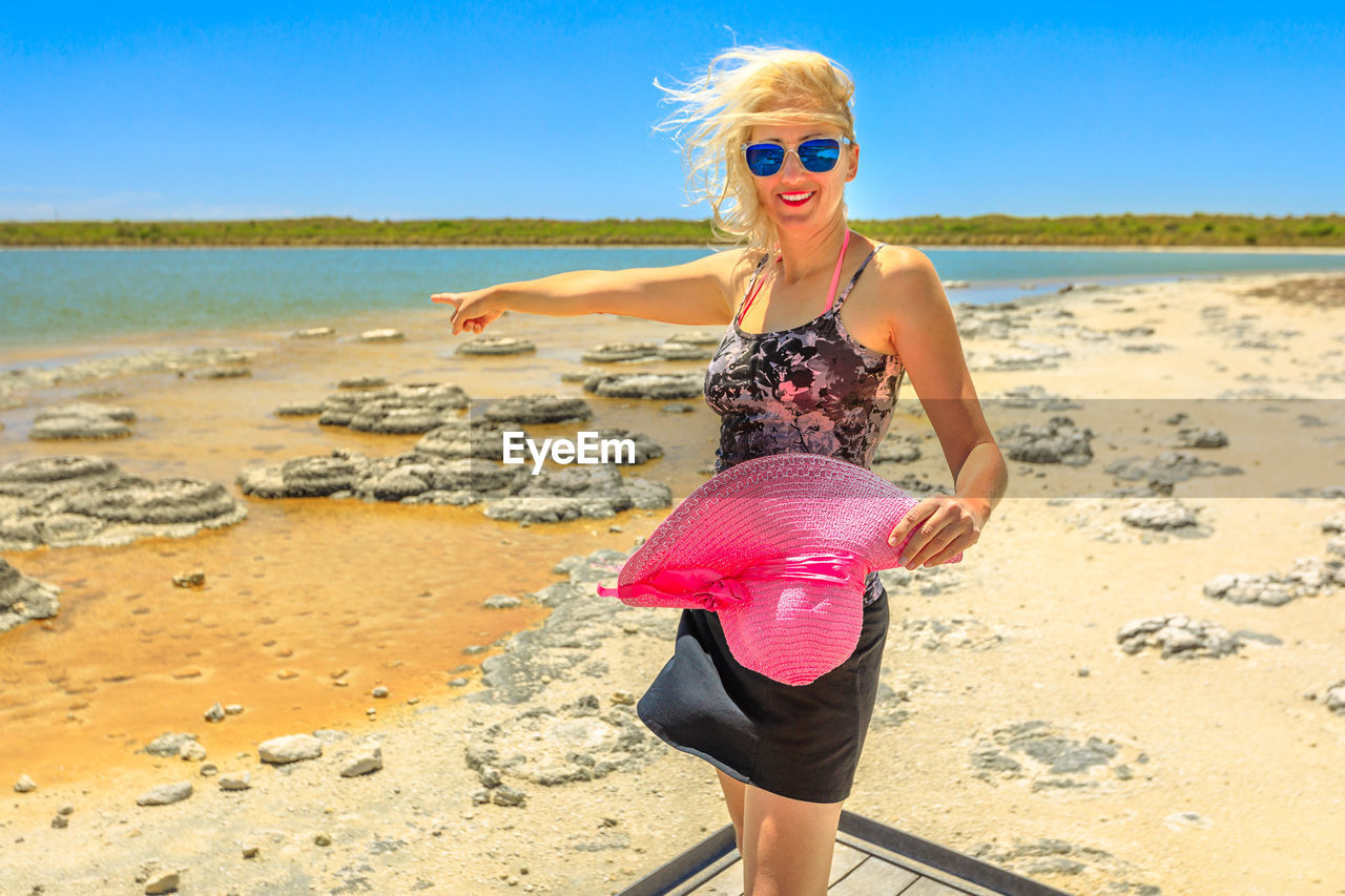 Full length portrait of woman pointing while standing on shore against sky