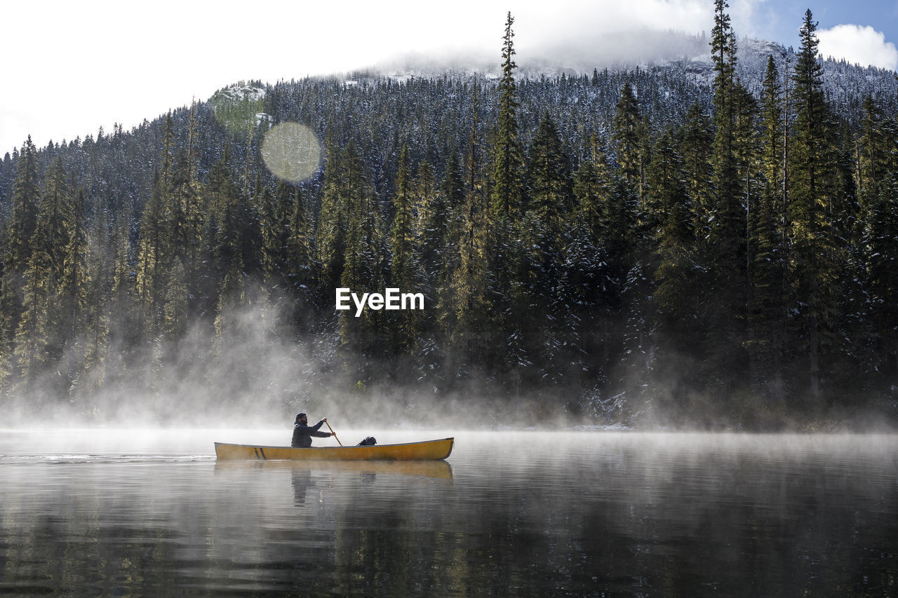 Man paddles canoe on lake with mist and fog on sunny day by forest