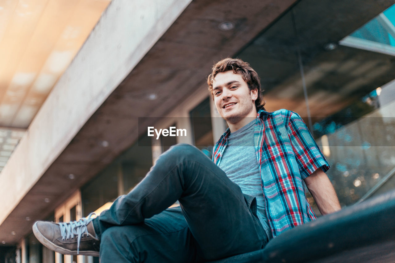 Low angle portrait of man smiling while sitting on seat against building