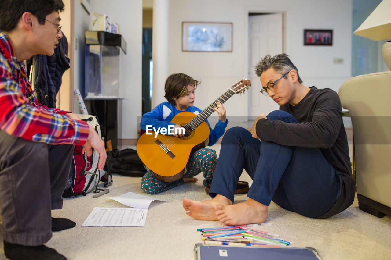 A small boy plays guitar for his father and uncle in living room