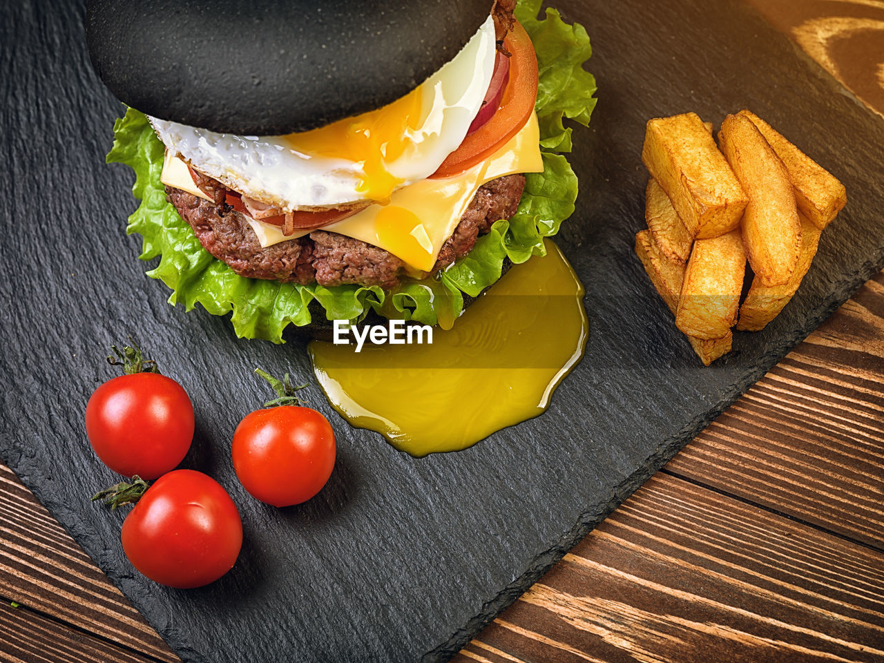 Black burger with an egg on a stone plate. cherry tomatoes. fried potatoes.