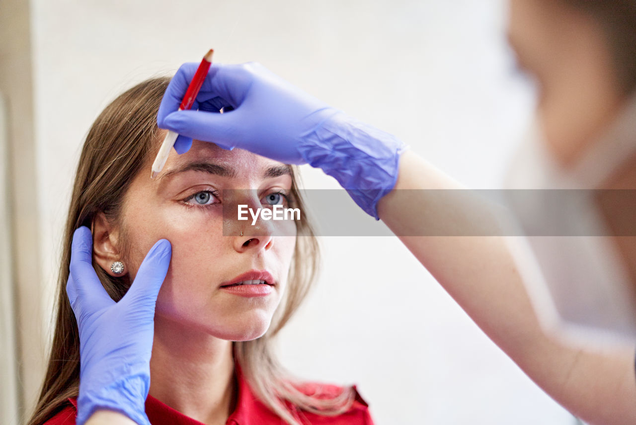 Blur beautician in latex gloves drawing line with white pencil while correcting shape of eyebrows of young woman during work in aesthetic clinic