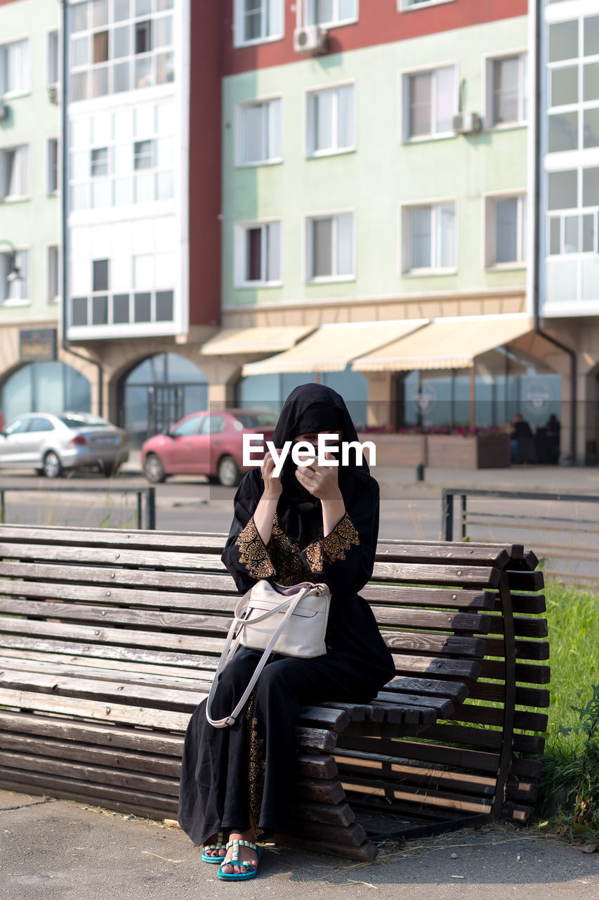 A muslim woman is resting a bench in the park, tidying herself up by looking in a mirror.