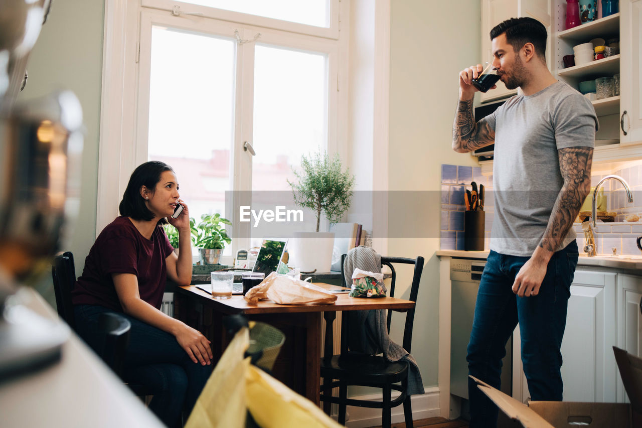 Woman using smart phone while man having drink at kitchen during relocation