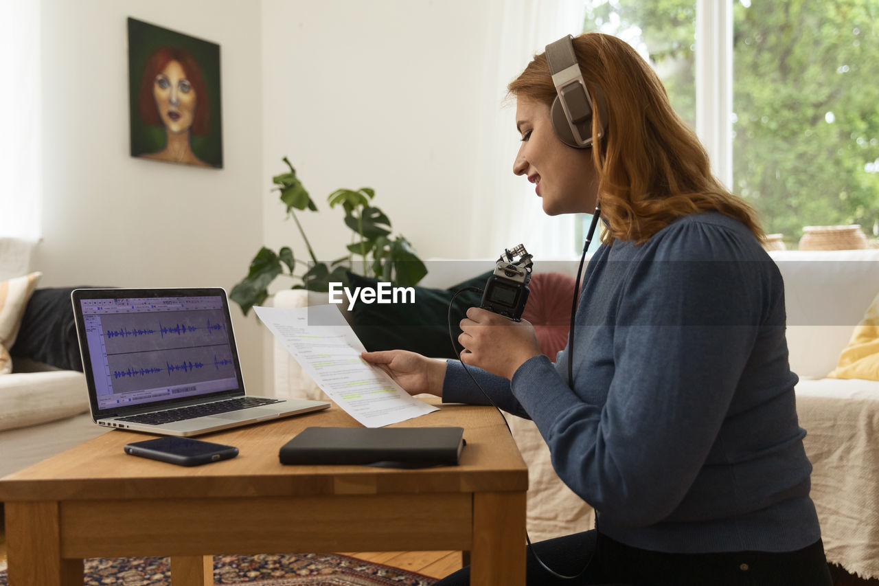 Delighted female radio host recording podcast while using mic and reading notes from paper at home