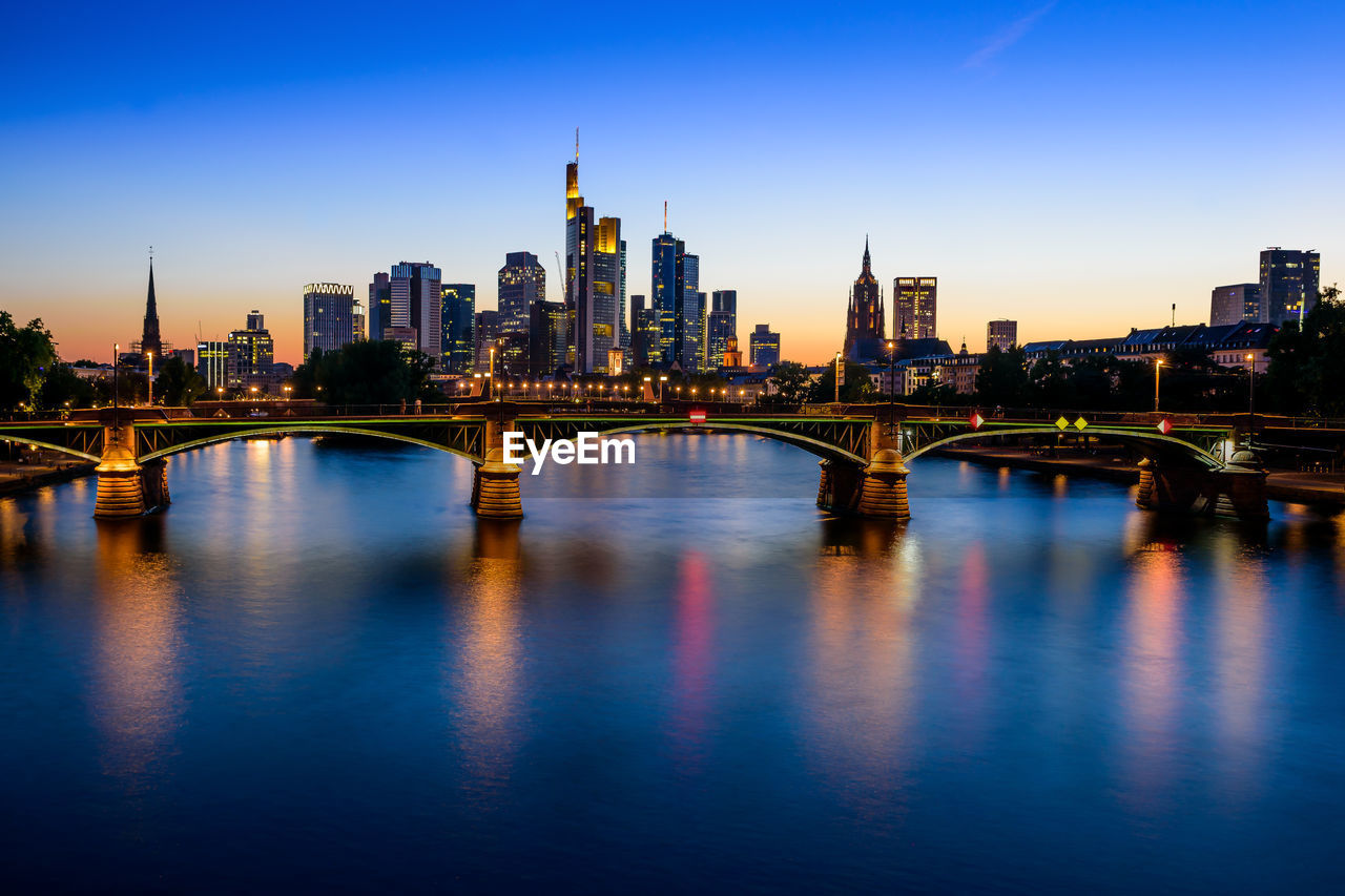 Illuminated bridge over river by buildings against sky in city