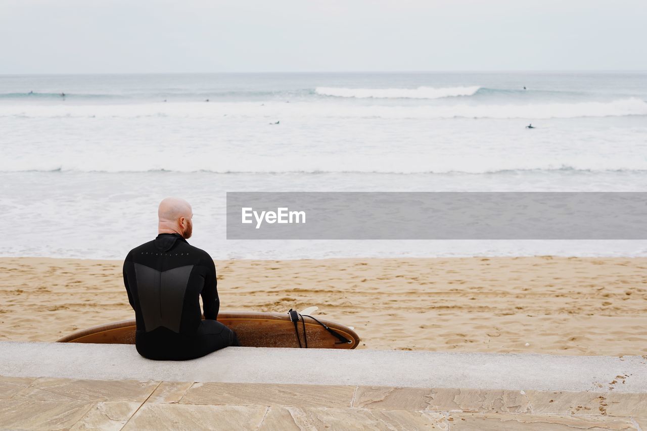 REAR VIEW OF MAN SITTING ON BEACH AGAINST SEA