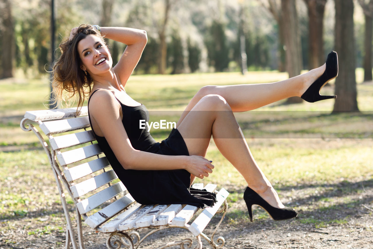 Side view of cheerful young woman sitting on bench at park