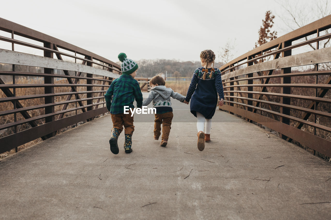 Three siblings holding hands running on bridge away from camera