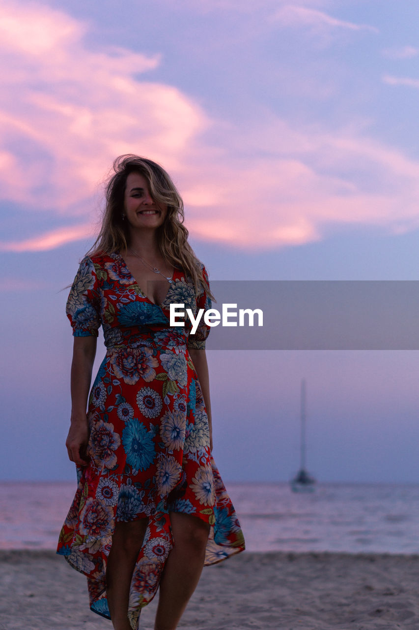 YOUNG WOMAN STANDING ON BEACH AGAINST SKY DURING SUNSET