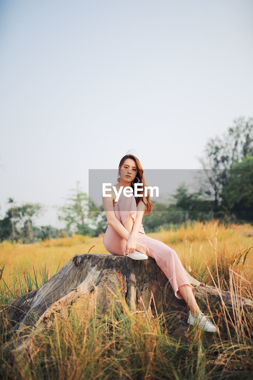 Portrait of young woman sitting on tree stump against clear sky