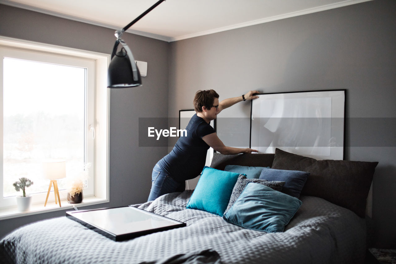 Pregnant woman arranging paintings on gray wall in bedroom at home