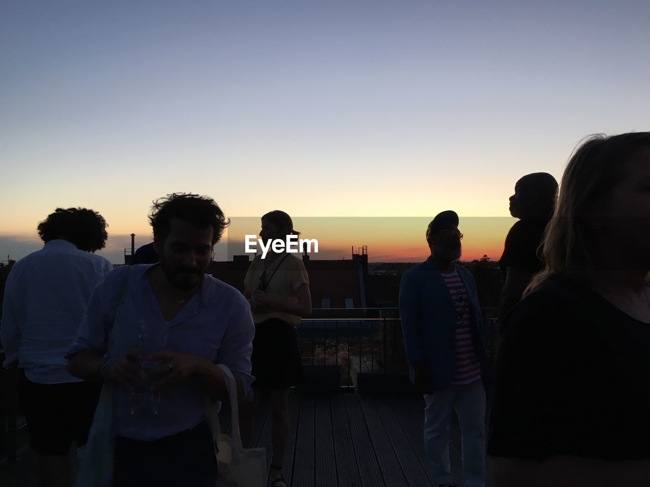 REAR VIEW OF PEOPLE SITTING ON SHORE AGAINST SKY DURING SUNSET