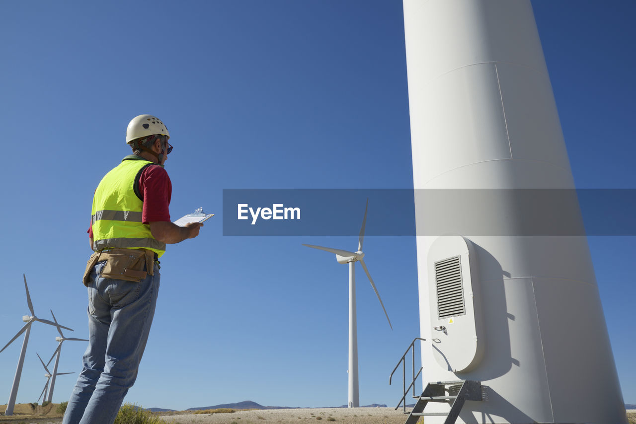 Low angle view of engineer holding clipboard while standing by windmills against sky