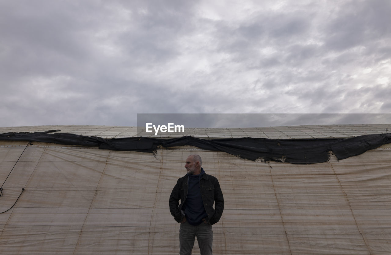 Adult man standing in front of plastic greenhouses against cloudy sky in almeria, spain