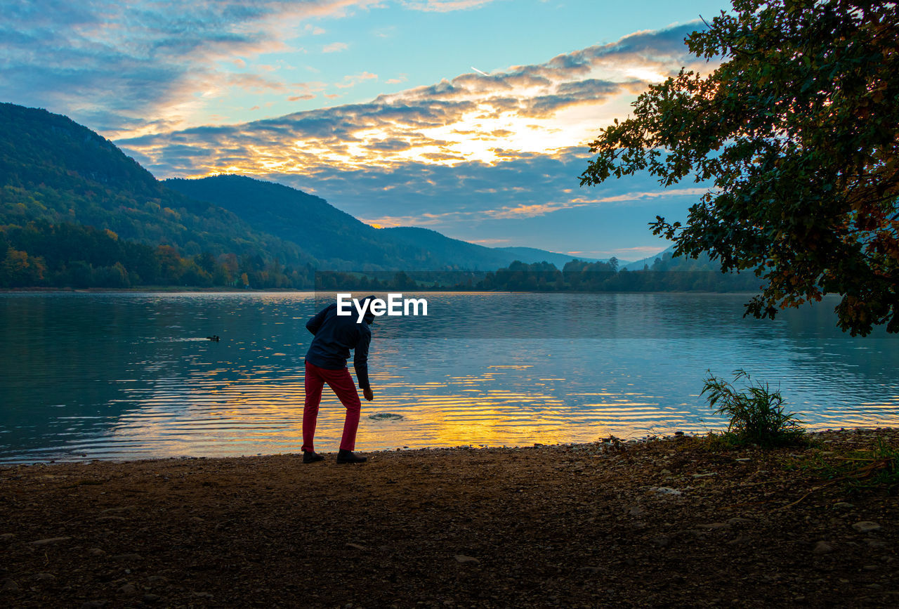 REAR VIEW OF MAN LOOKING AT LAKE AGAINST MOUNTAIN