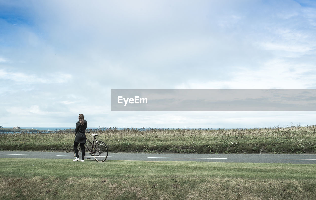 Rear view of woman with bicycle walking on road amidst field against cloudy sky