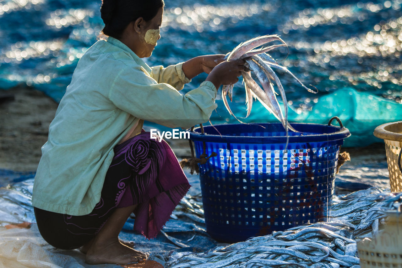 Side view of woman holding fish over basket at beach