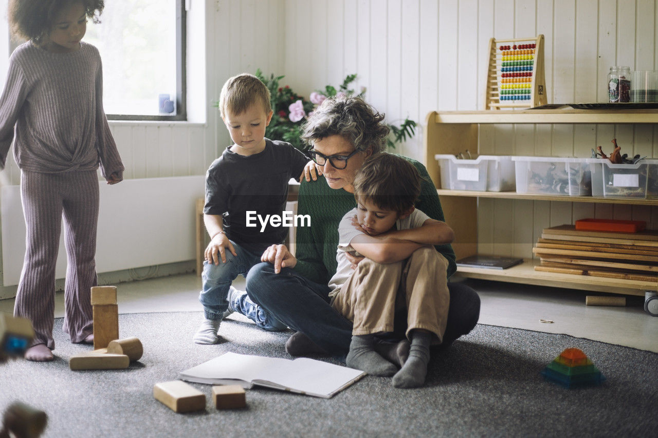 Group of kids sitting with female teacher reading book in classroom at kindergarten