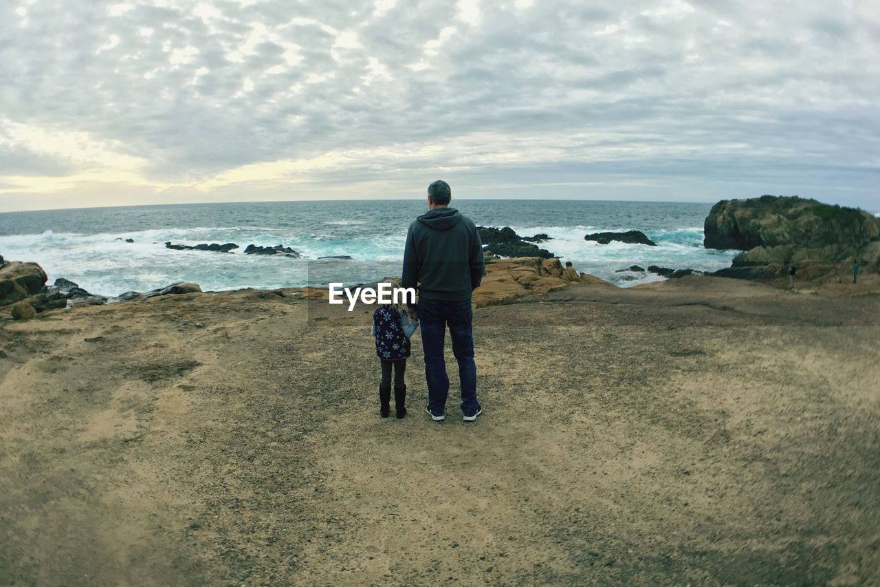 Full length rear view of father with daughter standing at sea shore against sky