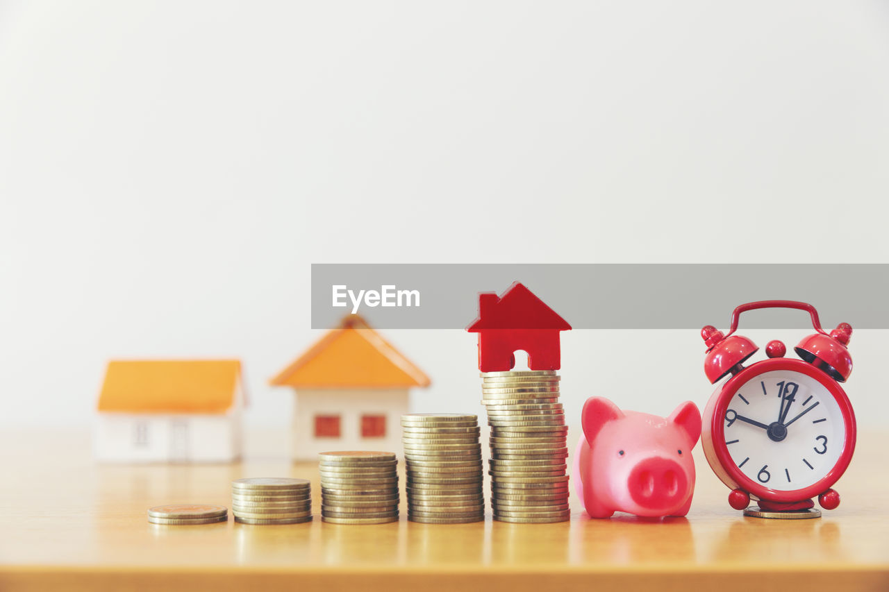 Close-up of coins with model homes and alarm clock on wooden table against wall