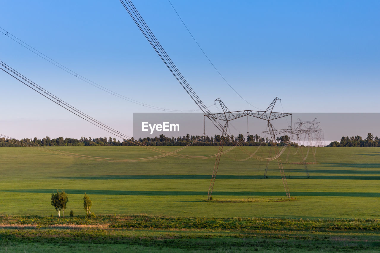 Scenic view of power line towers on green summer field against sky at day light