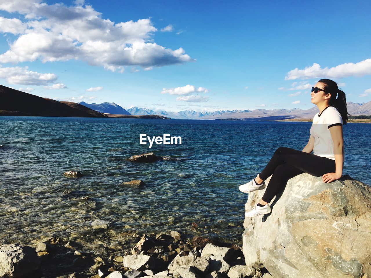 Young woman sitting on rock by lake against cloudy sky