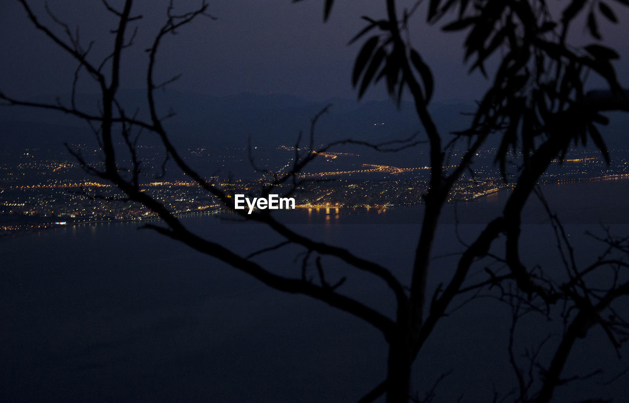 High angle view of sea and illuminated city against sky seen through silhouette tree