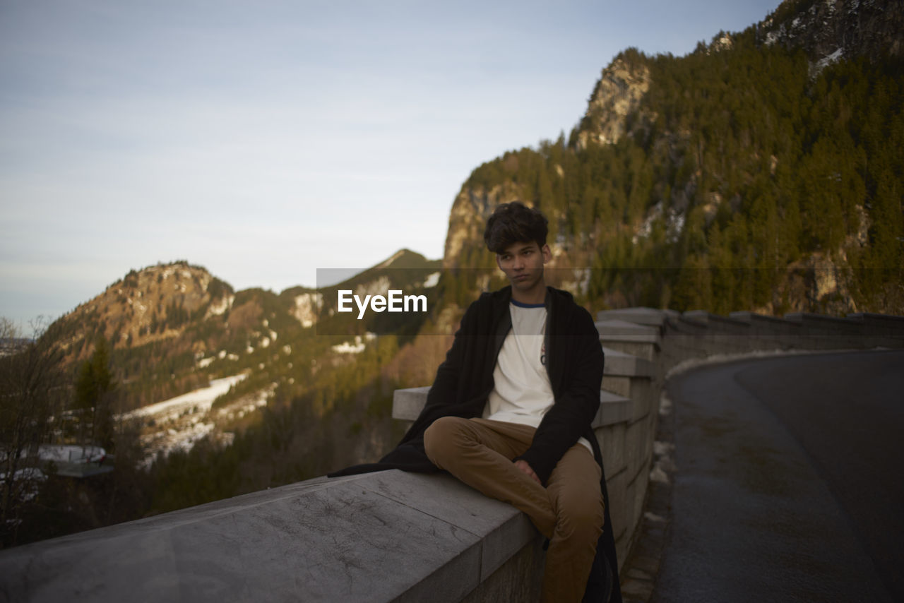 Man sitting on retaining wall against sky