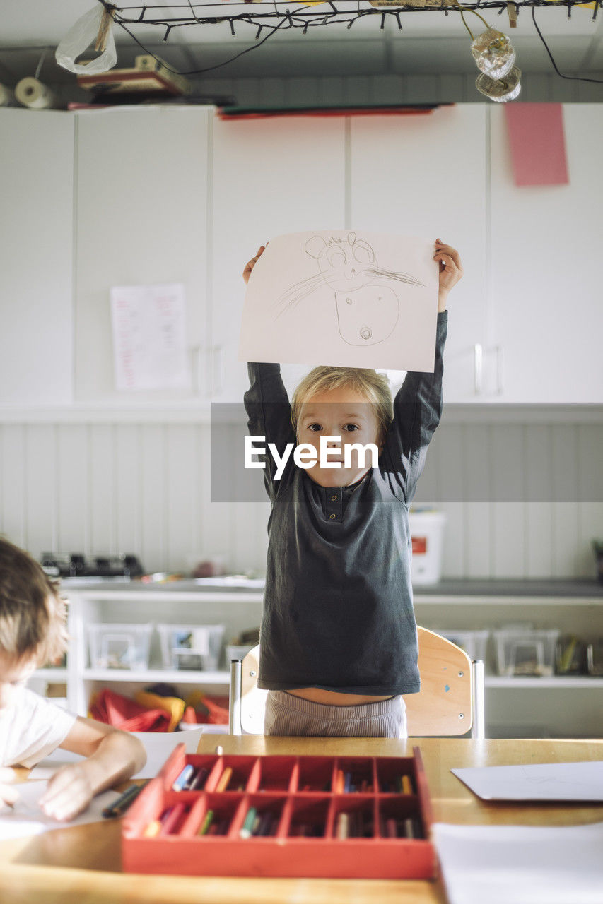 Portrait of girl showing paper with drawing while standing near bench in classroom