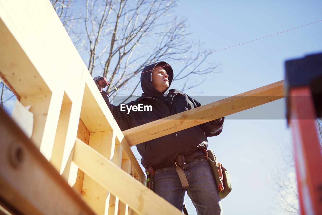 Low angle view of worker constructing wooden house against blue sky
