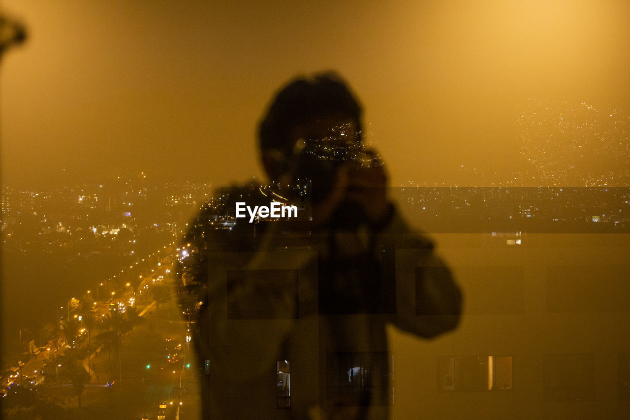 Reflection of man photographing on illuminated cityscape at night