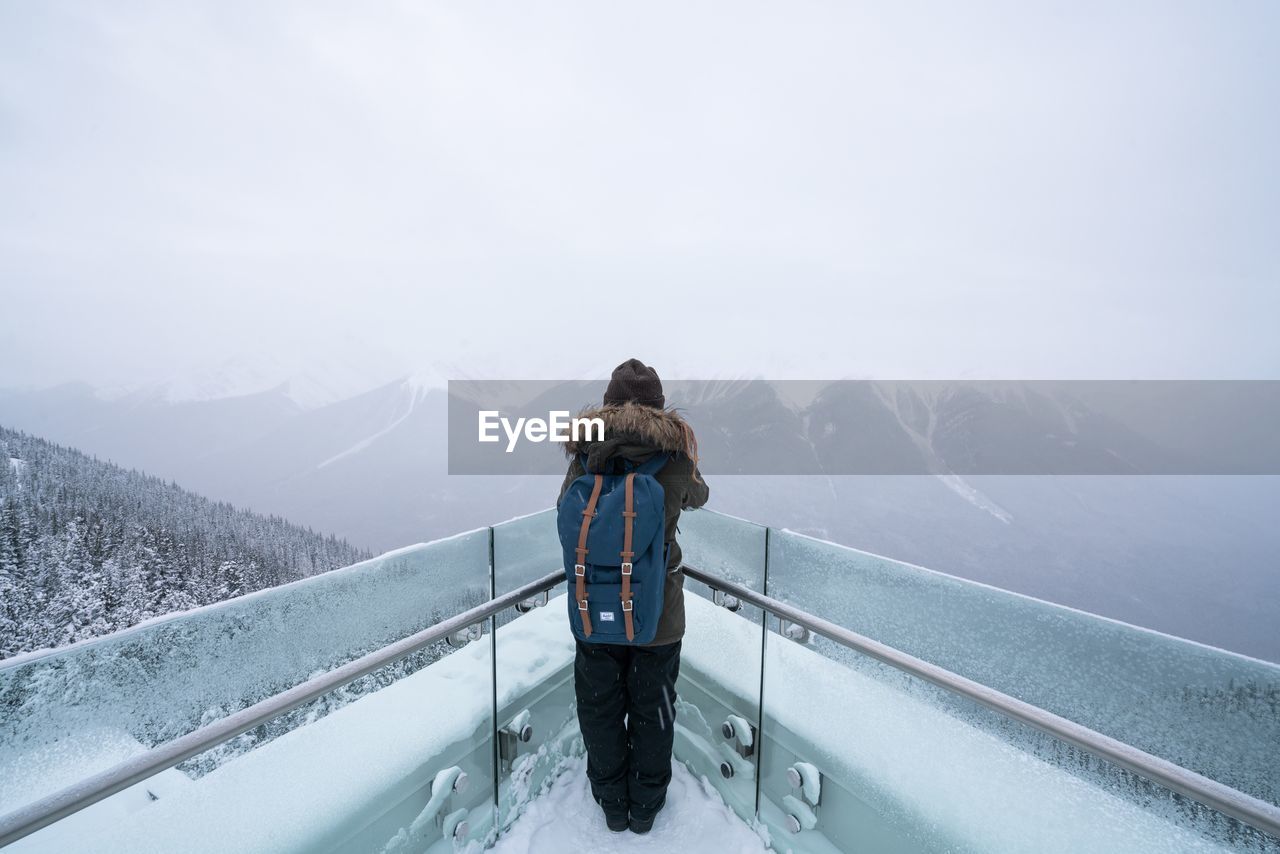 REAR VIEW OF MAN STANDING ON SNOW COVERED MOUNTAINS