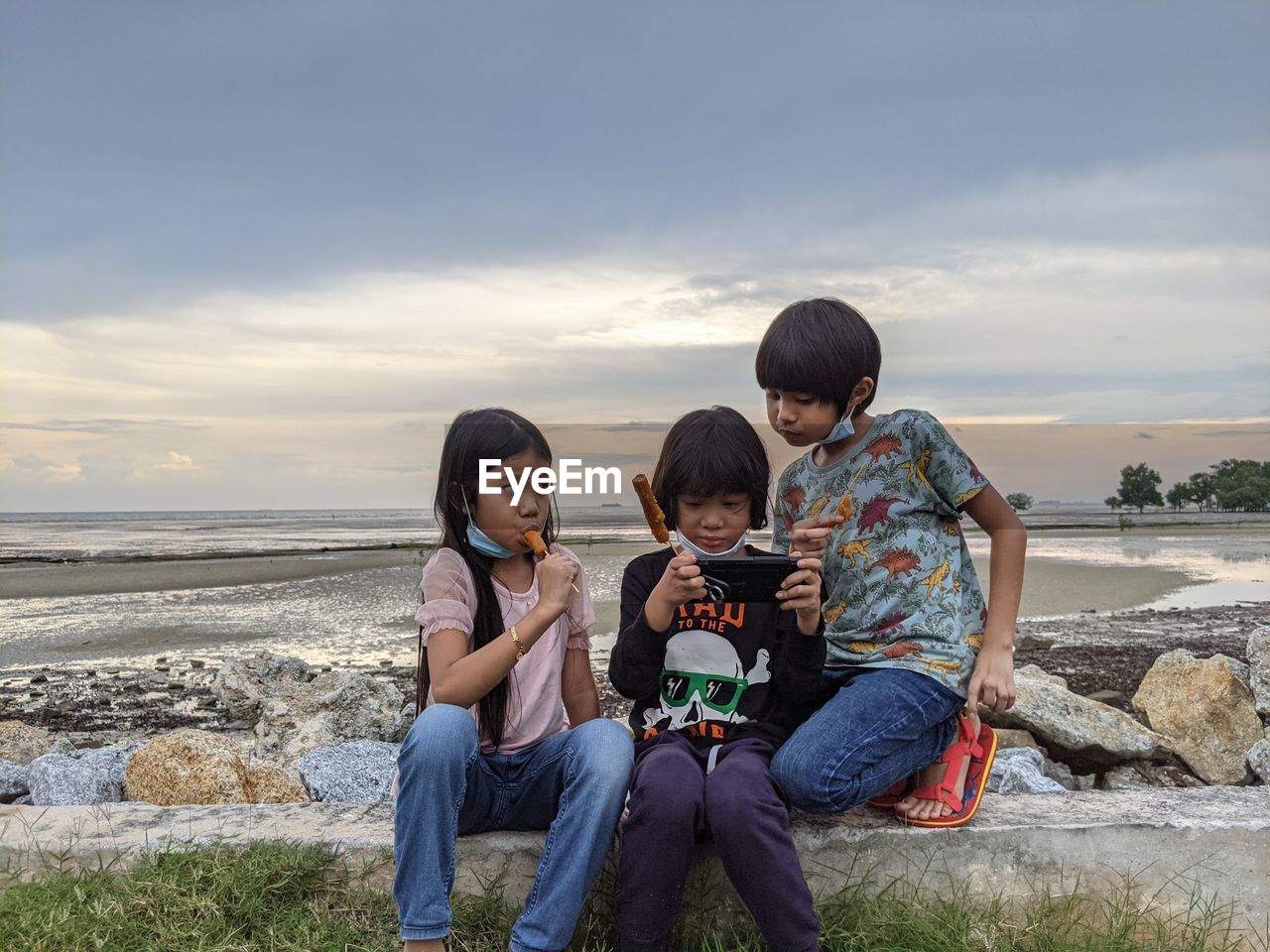 3 kids eating hotdog on stick, looking at smartphone isolated on beach background 