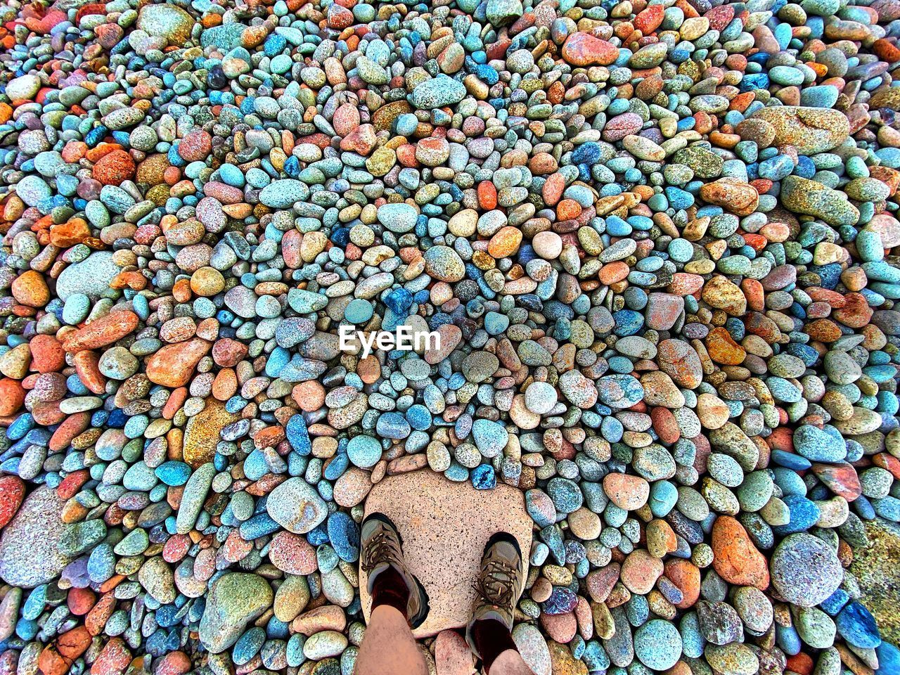 low section, human leg, pebble, shoe, one person, high angle view, gravel, abundance, day, standing, personal perspective, stone, directly above, soil, large group of objects, multi colored, flooring, outdoors, lifestyles, rock, human foot, full frame, pattern, nature, art, human limb, limb, sandal