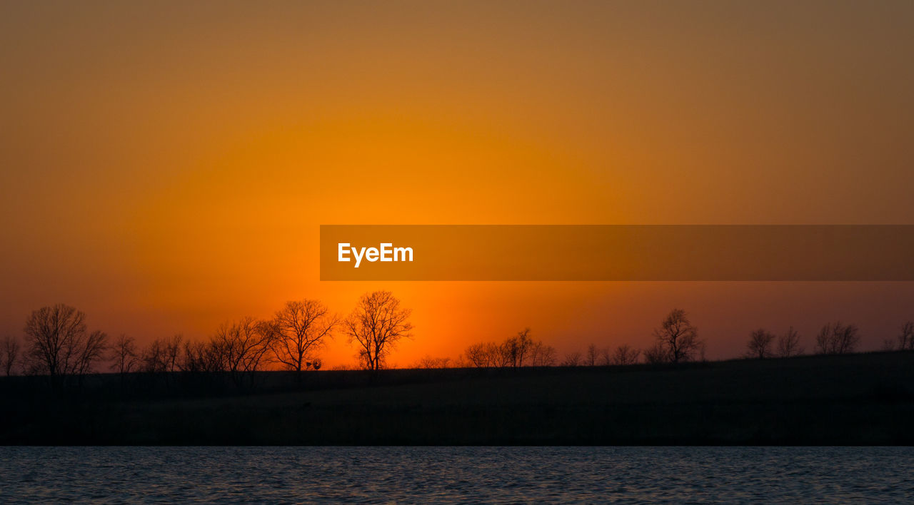 SILHOUETTE TREES BY LAKE AGAINST ORANGE SKY DURING SUNSET