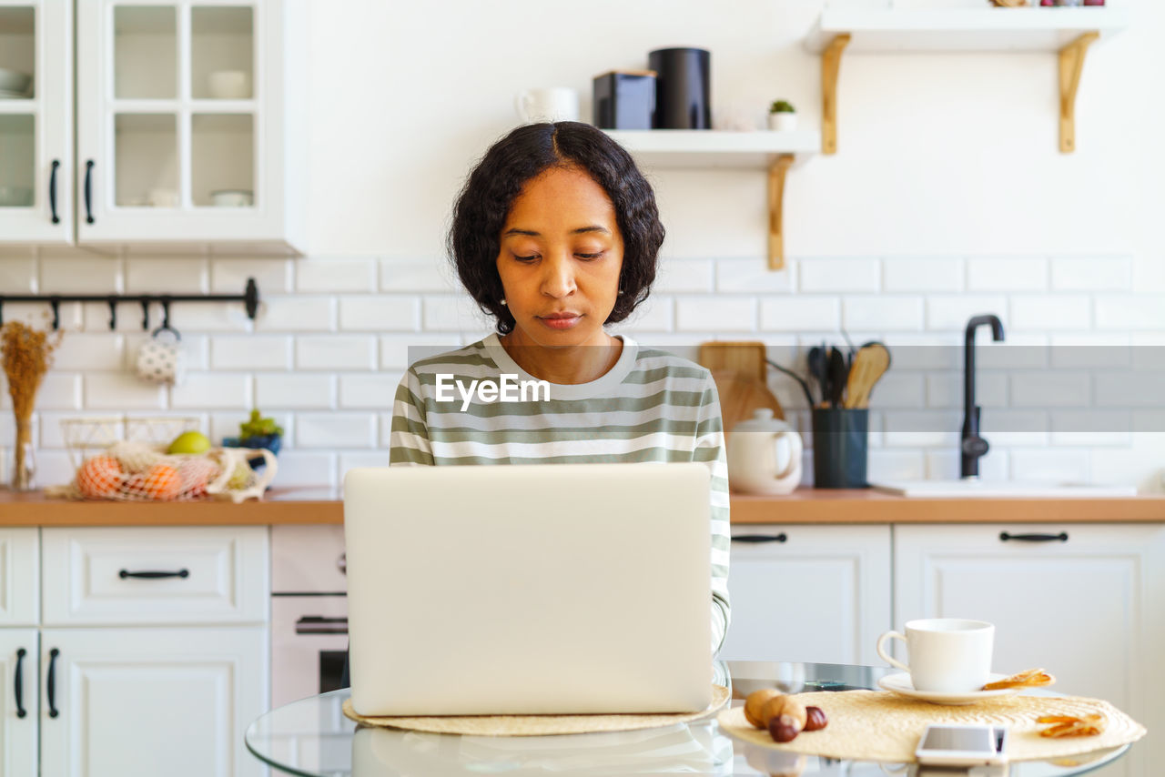 Focused african-american female working from kitchen on laptop. concept of home remote office