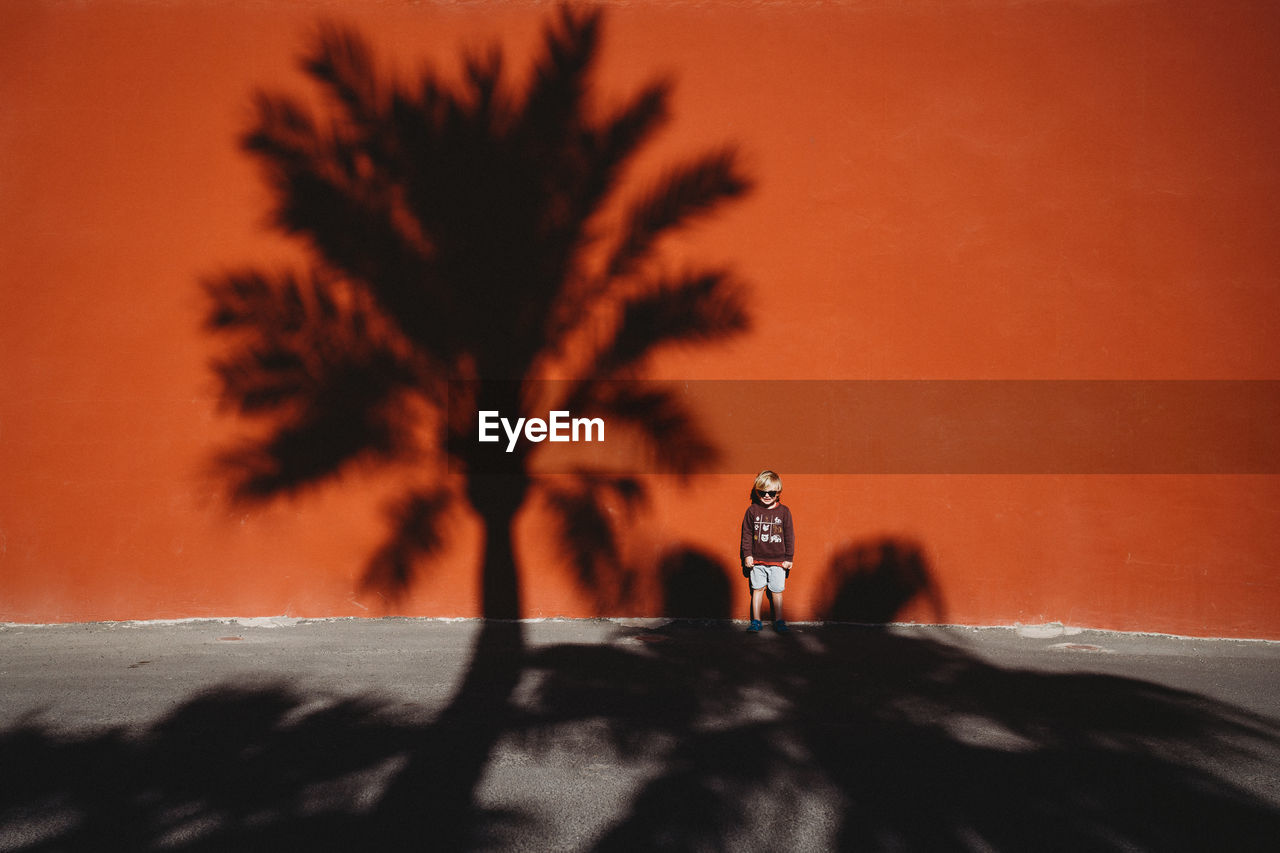 Boy standing against a red wall with the shadow of a palm tree