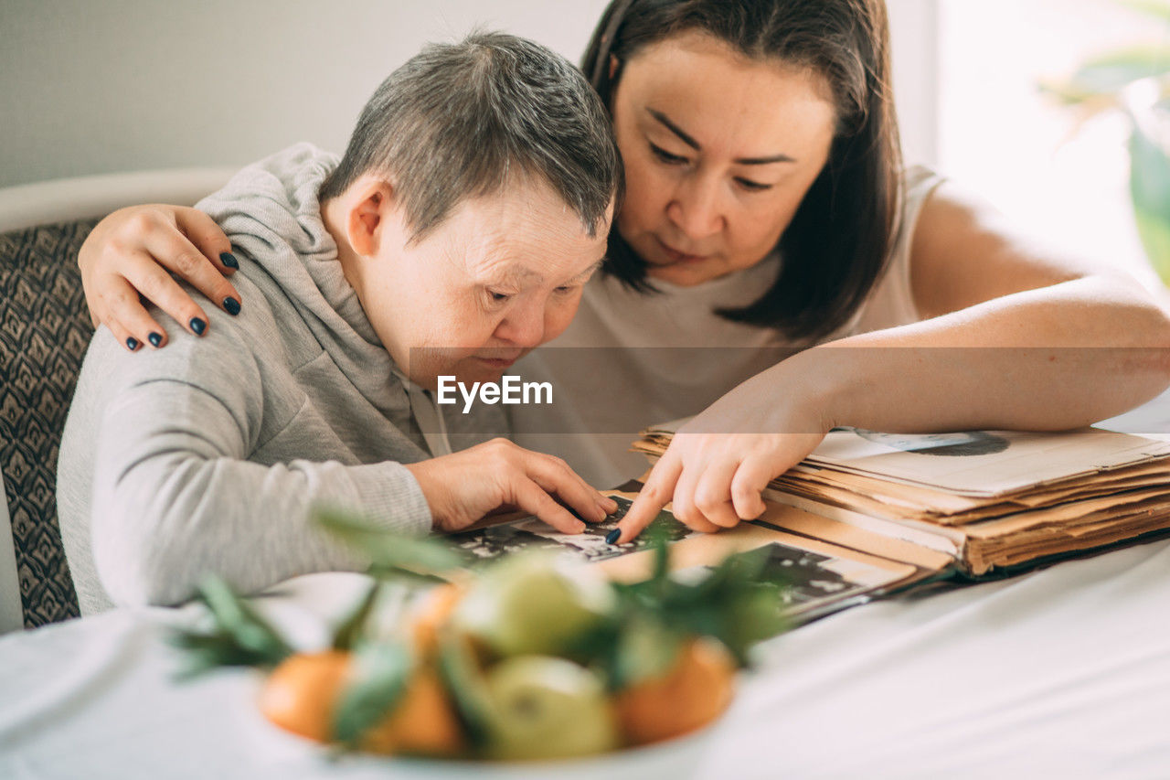 Asian girl points a smiling elderly woman with down syndrome to a photo in an old album.