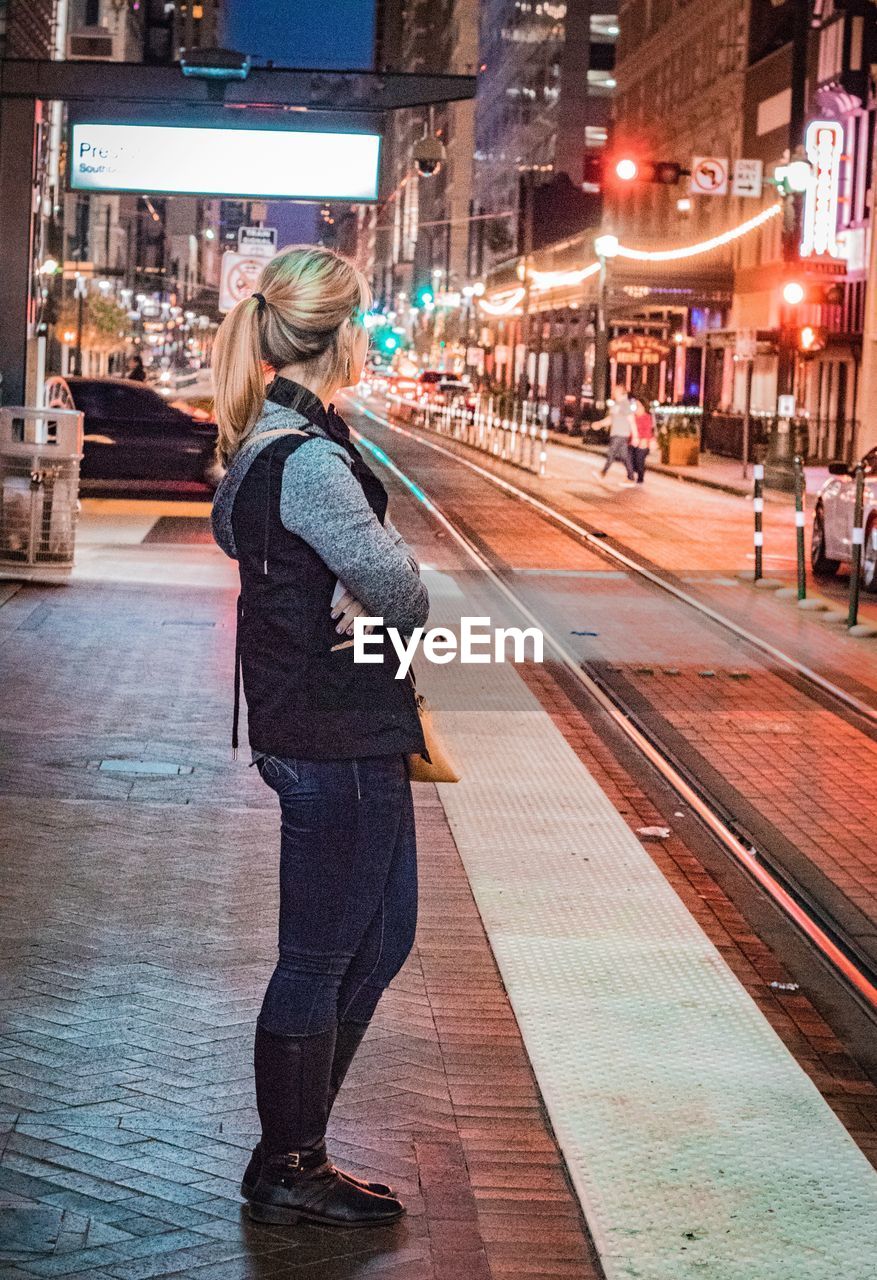 FULL LENGTH REAR VIEW OF WOMAN STANDING IN ILLUMINATED CITY